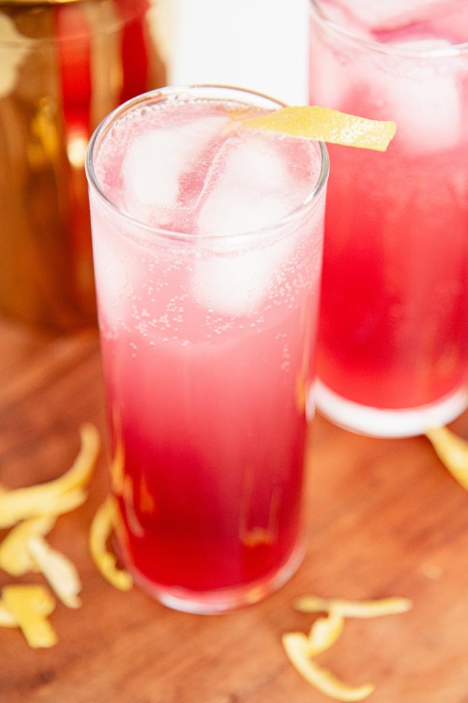 an over head picture of a hard seltzer with a lemon twist on top, and lemon zest around the glass, you can see part of another seltzer and a gold cocktail shaker behind it 