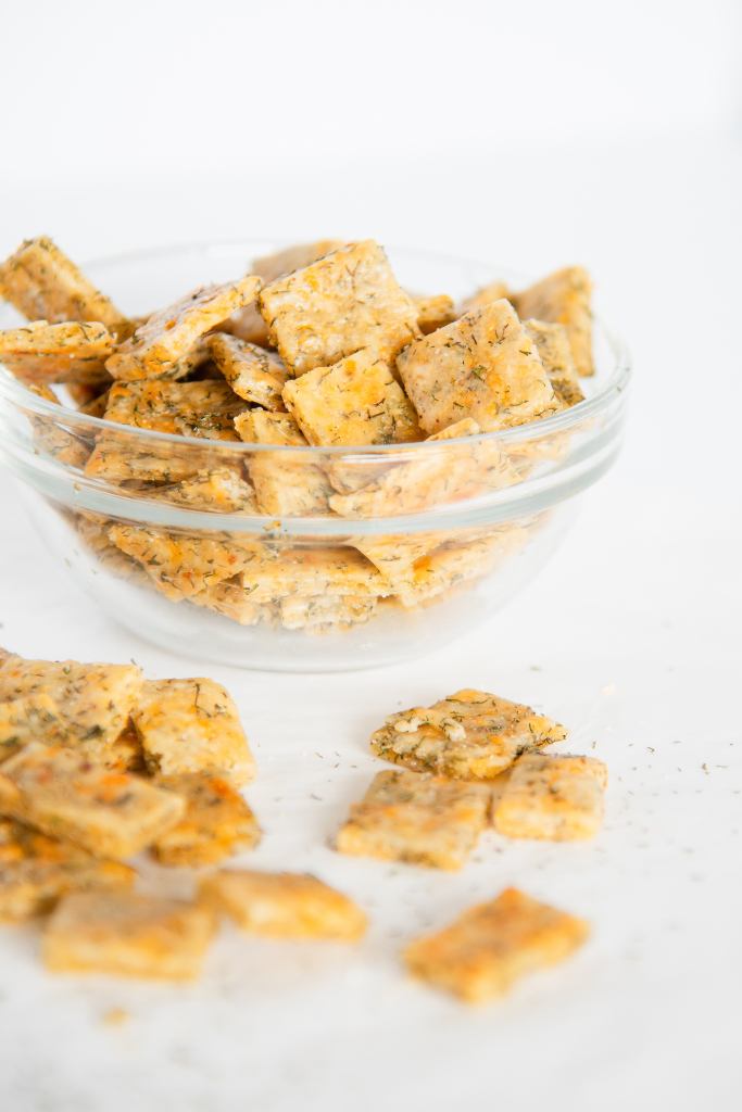 a small glass bowl of crackers, with some crackers in front of it on the table