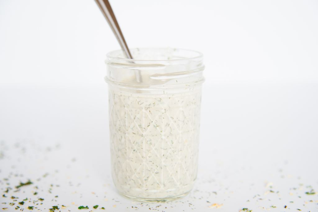 a picture from the side of a mason jar of ranch dressing with a spoon in it, and some spilled dill weed around it