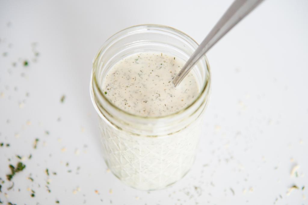 an overhead picture of a jar of ranch dressing with a metal spoon in it, there is some dill weed spilled around the jar