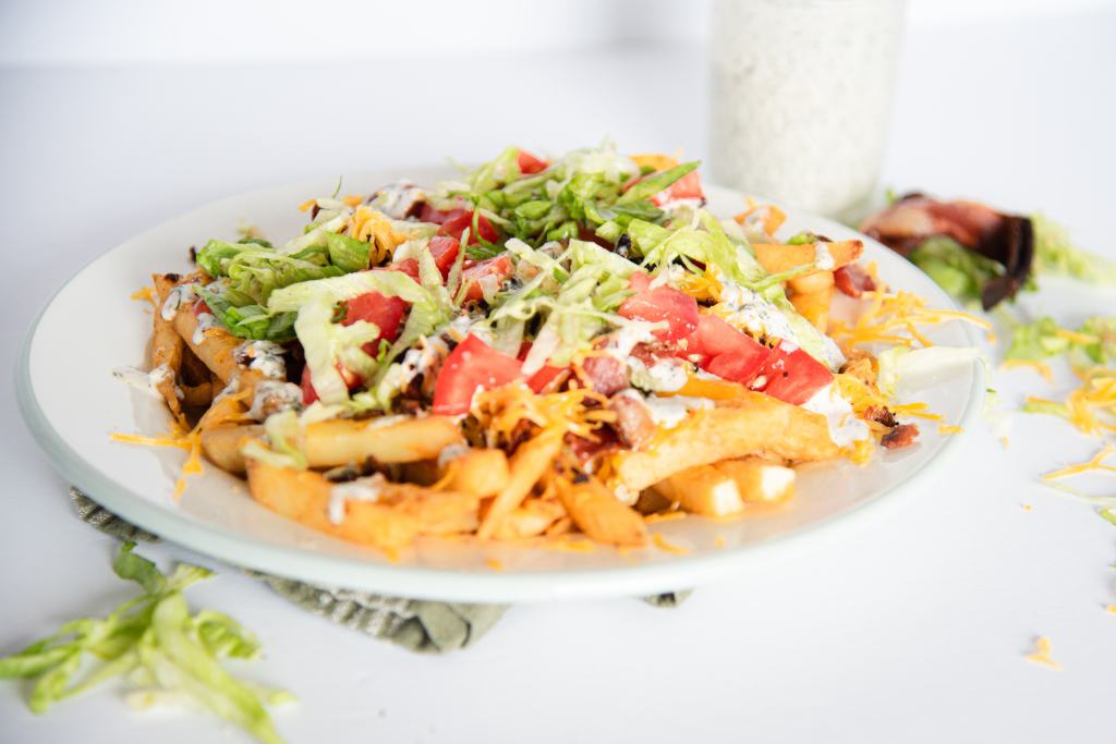 a plate of loaded fries with some shredded cheese and lettuce next to the plate, and jar of ranch dressing and some bacon behind the plate 