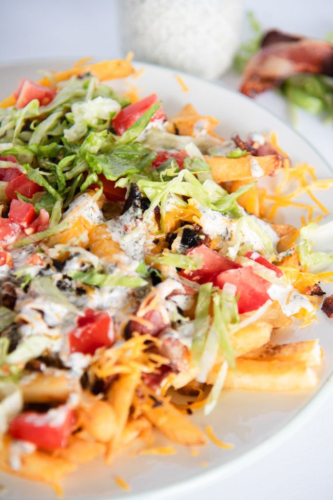 an overhead shot of a plate of loaded fries, there is a jar of ranch dressing and some lettuce and bacon behind the plate