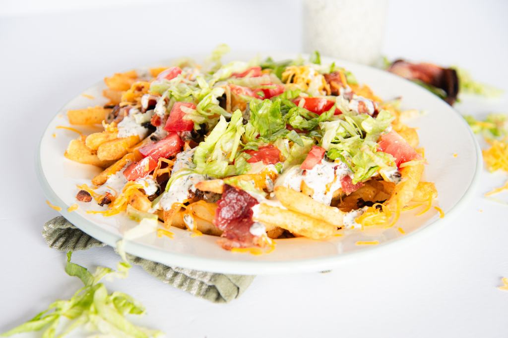 a plate of loaded cheddar bacon fries, there is some lettuce and  cheese around the plate, and some ranch dressing and a piece of bacon behind the plate 