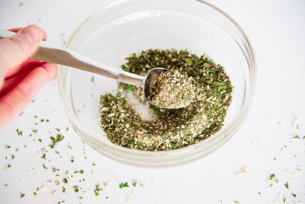 someone holding a measuring spoon of spices over a small bowl of spices