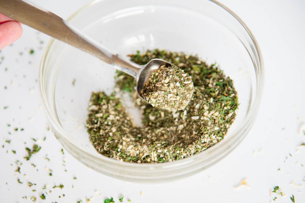 a measuring spoon of spices being hold over a glass bowl of spices