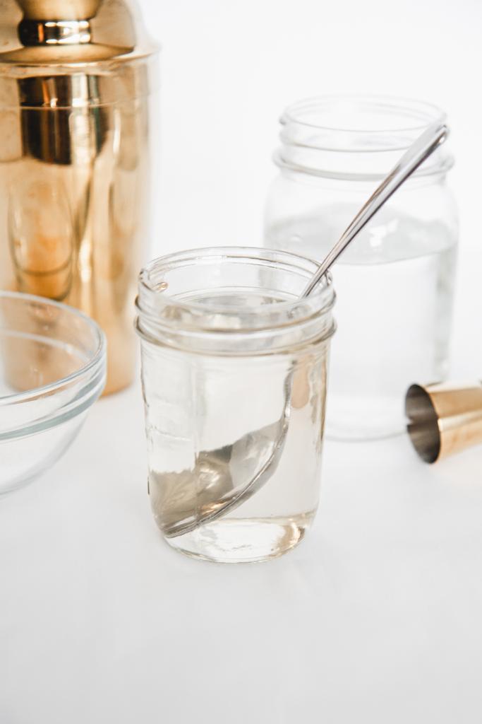 a jar of simple syrup with a spoon in it, there is a jar of water and a cocktail shaker behind it