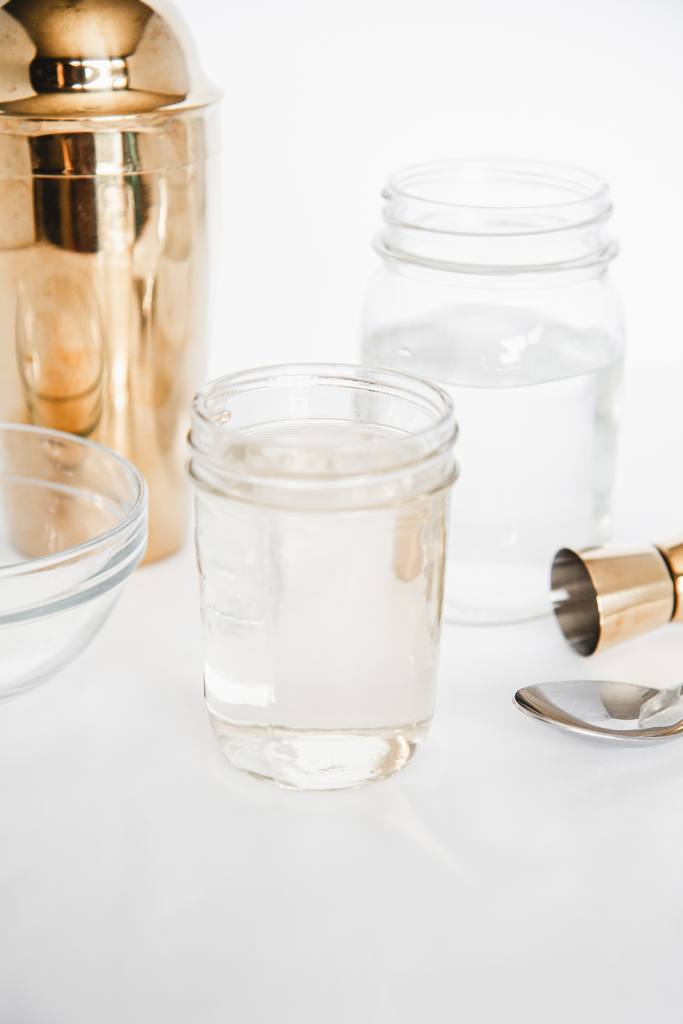 a jar of simple syrup with a jar of water, a bowl of sugar, a spoon, cocktail shaker and shot glass around it