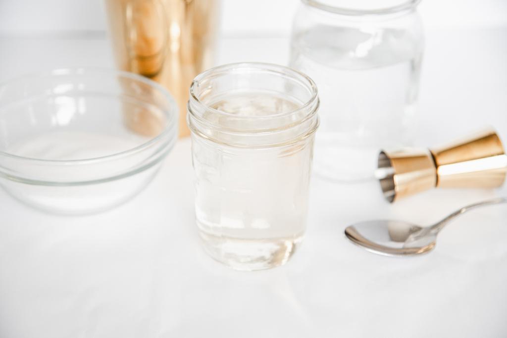 a jar of simple syrup with cocktail making supplies, a jar of water, a bowl of sugar and spoon by it 