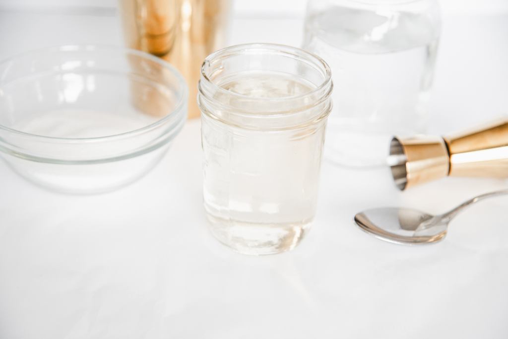 a jar of simple syrup, with a jar of water, a bowl of sugar, a spoon, a shot measuring cup and a cocktail shaker around it