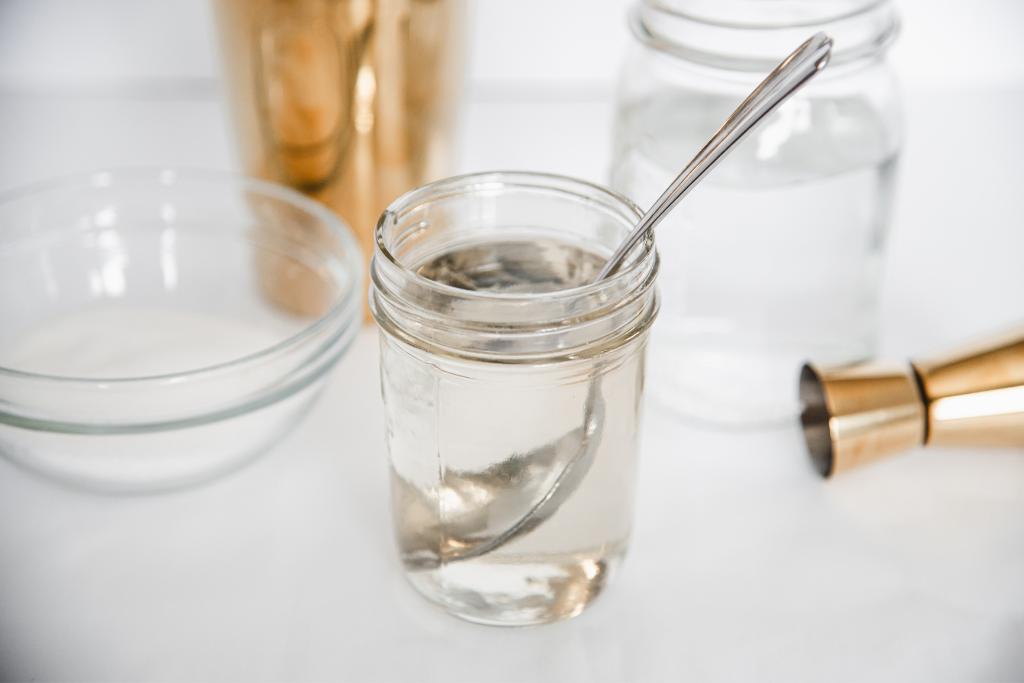 a jar of simple syrup with a mental spoon in it, there are ingredients and cocktail making supplies around it