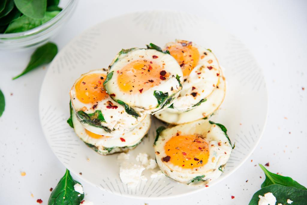 a stack of egg muffins on a plate, there is another muffin in front of the stack, and some crumbled feta cheese, there is a small bowl of spinach to the left side of the plate 