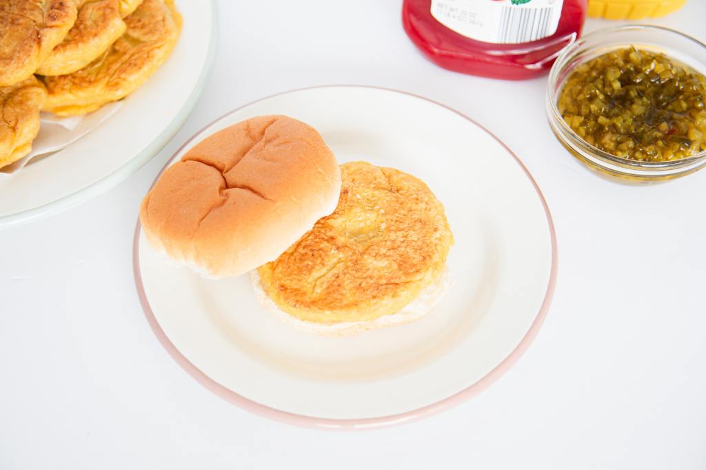 a chickpea burger patty on a bun, the top of the bun is resting it, there is a plate of burgers and some burger toppings next to it