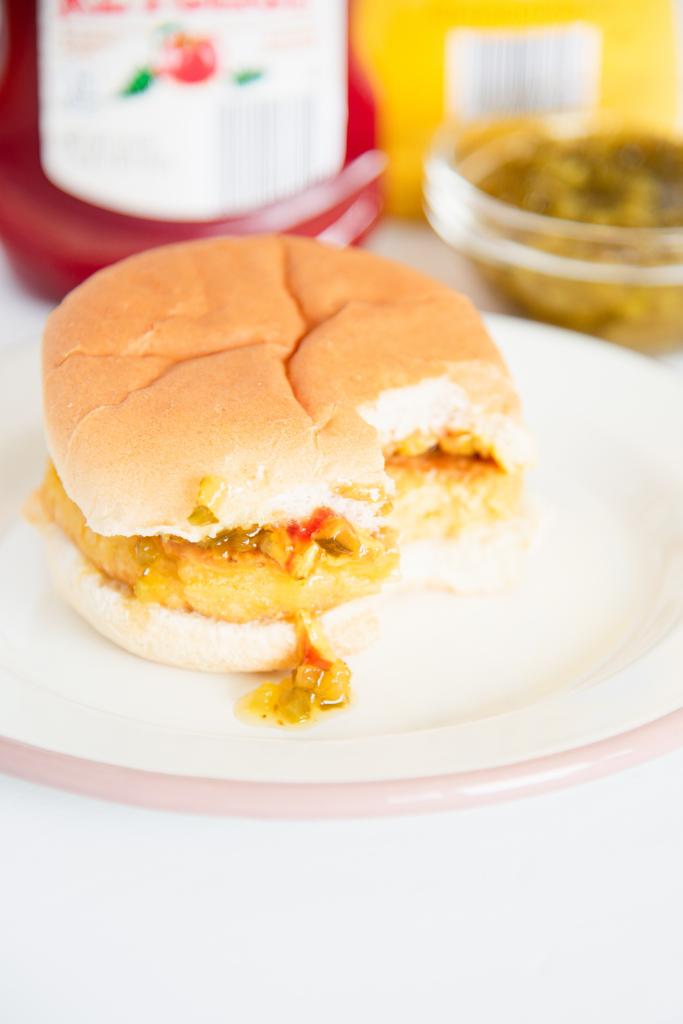 a finished chickpea veggie burger on bun with toppings, there is a bite taken out of it, and some toppings dripping down the side. You can also see some bottles and a bowl with toppings behind the black 