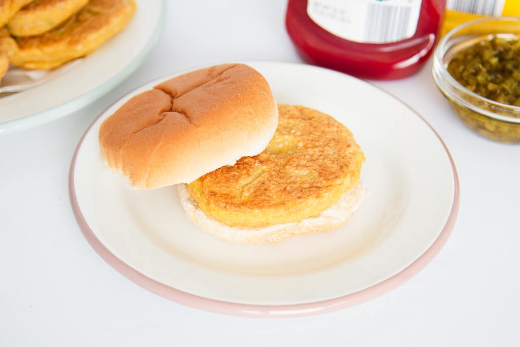 a chickpea burger patty on a bun, the top bun is resting on it, there is plate with more patties and some toppings on either side of the plate