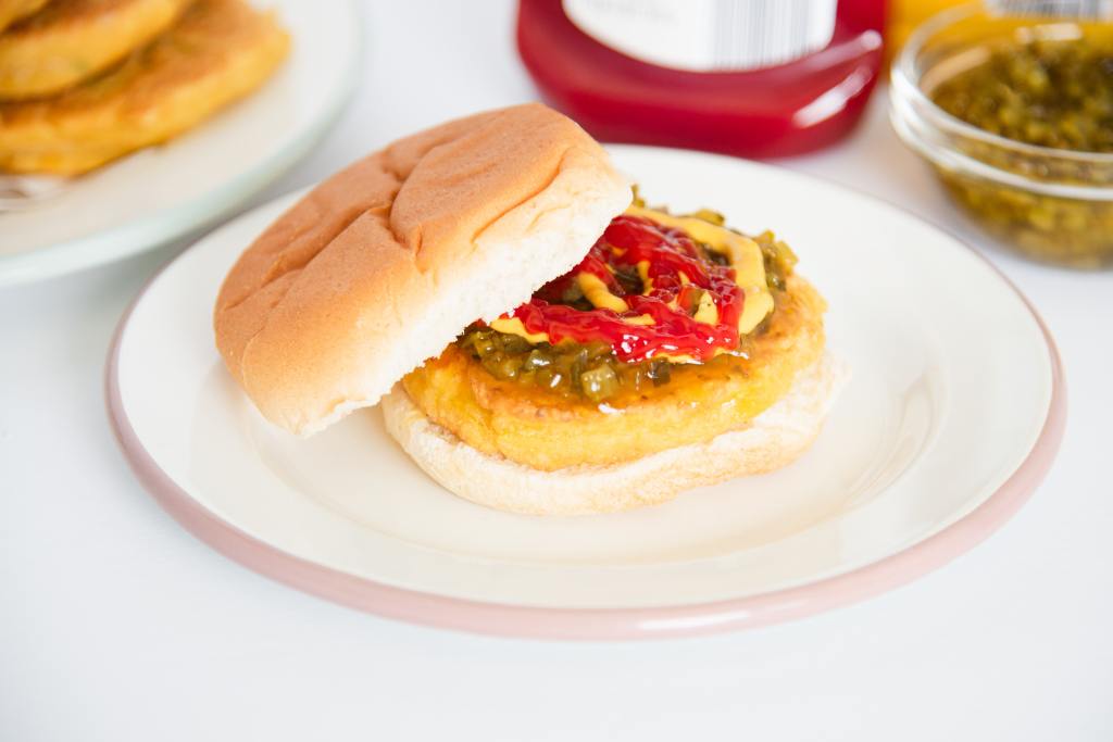 a chickpea burger on a hamburger bun with toppings on it. There is a plate of burgers and some toppings behind it