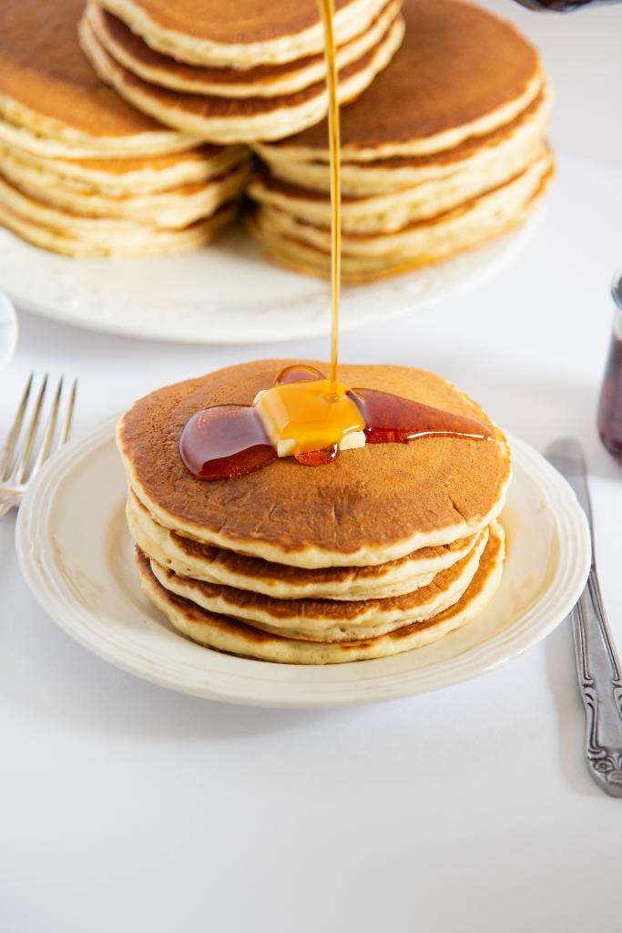 syrup being poured into a stack of pancakes, there is also some butter on the pancakes, and you can see another plate of pancakes behind the stack