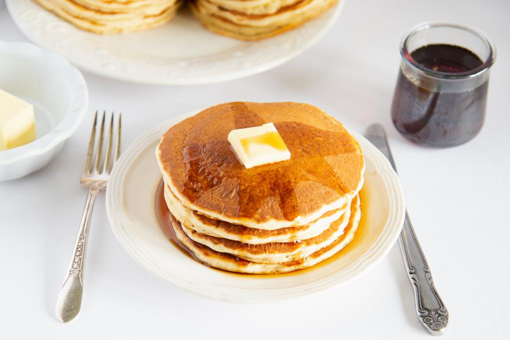 a stack of pancakes on a plate with butter and syrup, there is a fork and a knife of the side of the plate, you can also see a dish of butter and a jar of syrup