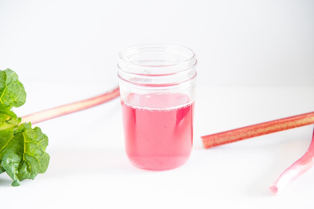 a jar of rhubarb syrup with three stalks of rhubarb around it, you can see part of a leaf on the top of one stalk