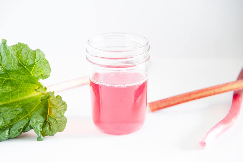 a small glass jar of rhubarb syrup with three stalks of fresh rhubarb around it