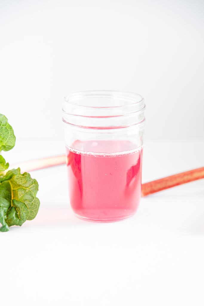 a close up of a jar of rhubarb syrup, you can see part of rhubarb stalk and a rhubarb leaf next to it