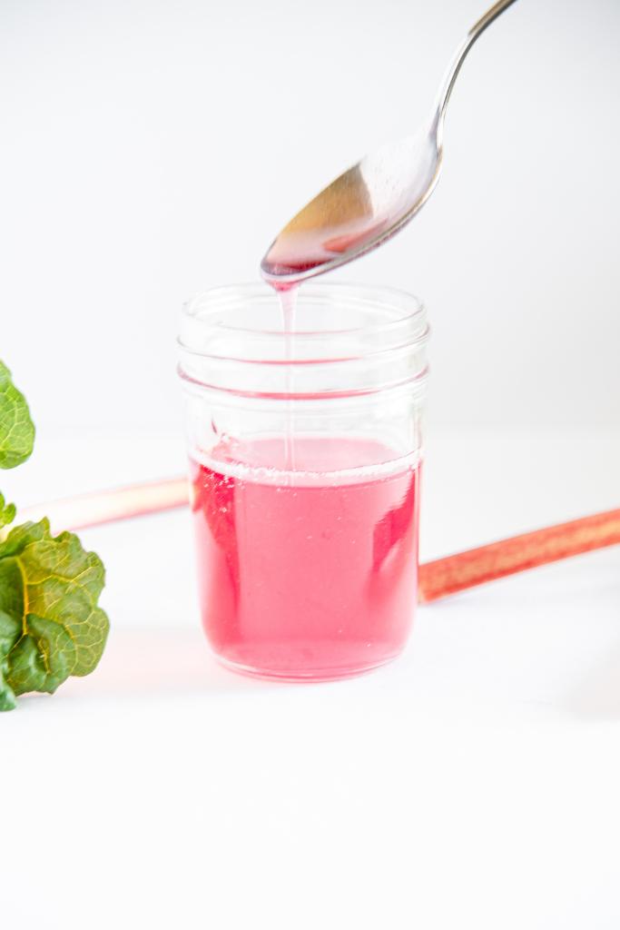 a metal spoon being lifted out of a jar of rhubarb syrup, there is syrup running off the spoon back into the jar