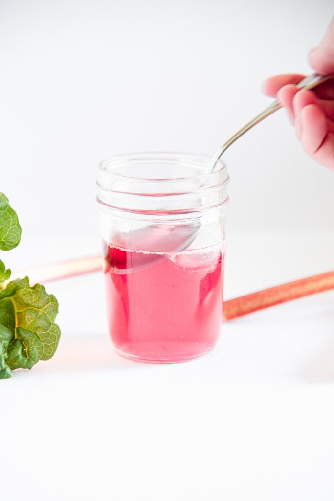 someone dipping a metal spoon into a jar of rhubarb syrup, there is a rhubarb stalk and leaf next to the jar