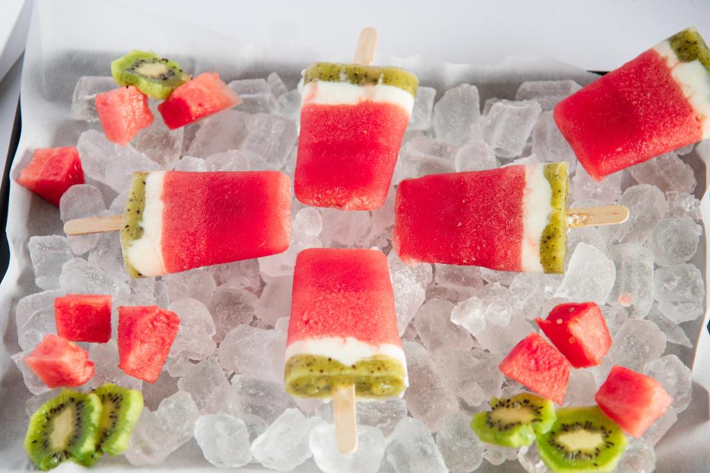 four watermelon popsicles set in a start shape on a tray of ice, there is another popsicle to the right side, and some fresh watermelon and kiwi around them