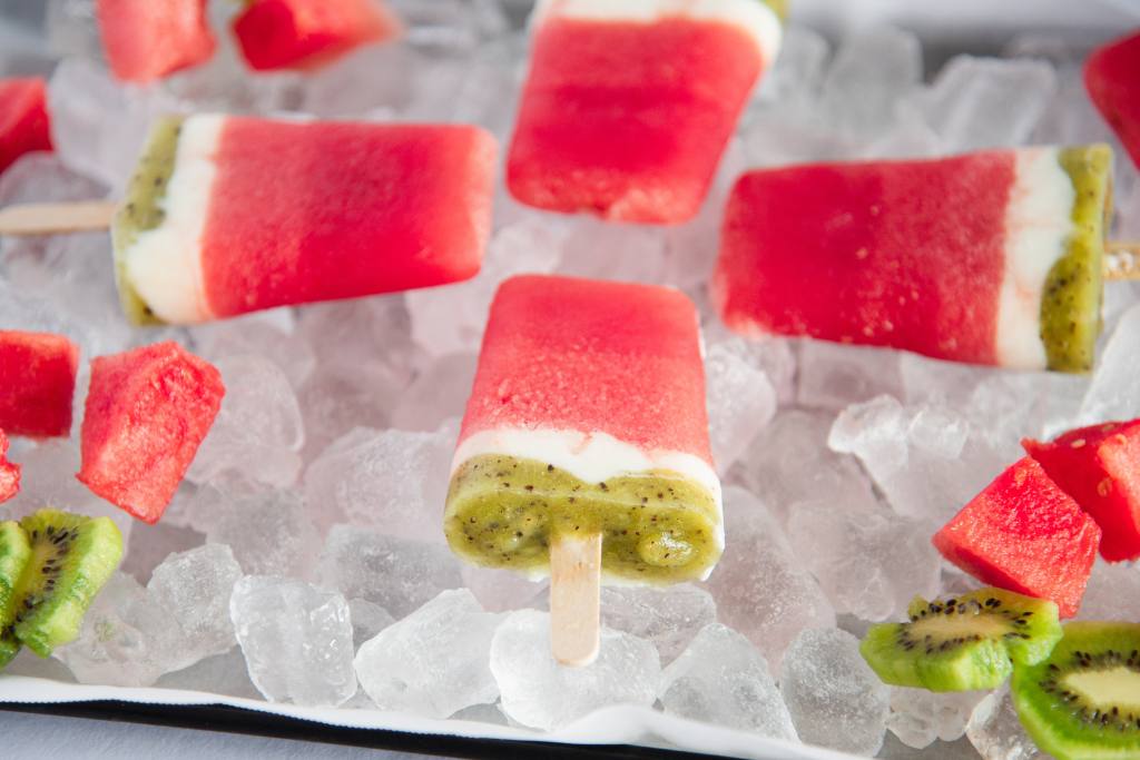 a close up of bottom of a watermelon Popsicle, ton a tray of ice, there are three other popsicles on the tray, and some watermelon chucks and kiwi slices  