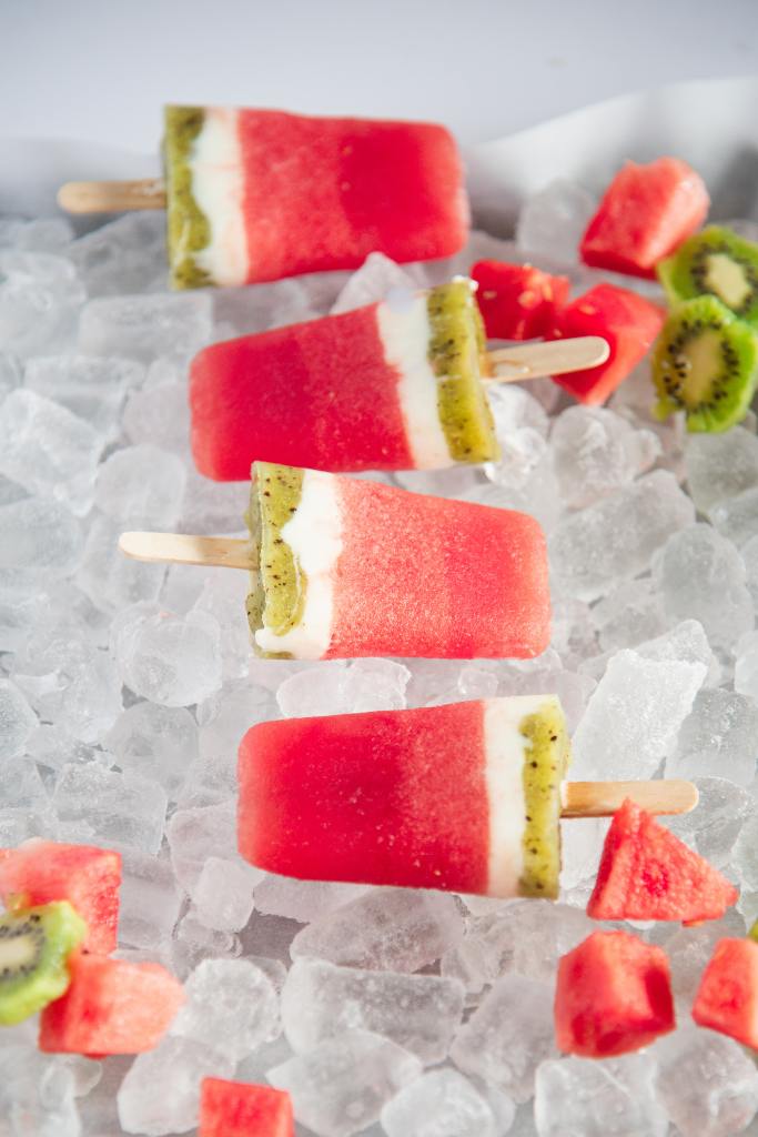 four watermelon popsicles on a tray on ice, there are some watermelon chucks and sliced kiwi around them