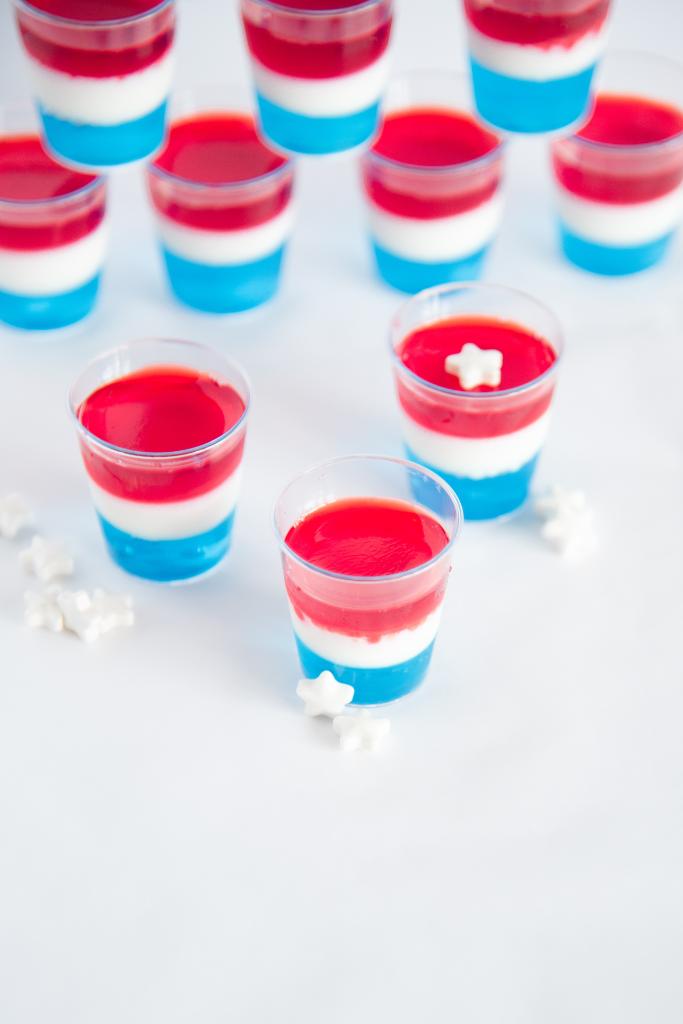 3 layered jello shots in front of a stack of jello shots, there are some white star sprinkles around the shots 