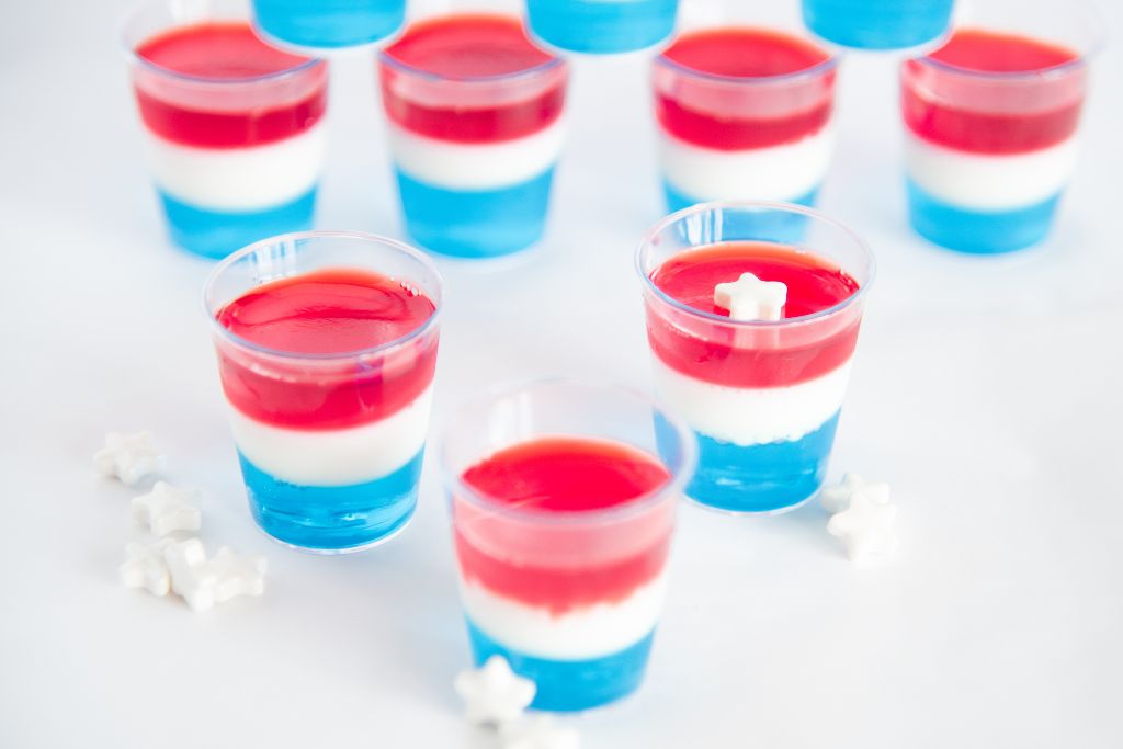 three jello shots in front of a stack of jello shots, there are white star sprinkles around them, and one shot has a star on top of it 