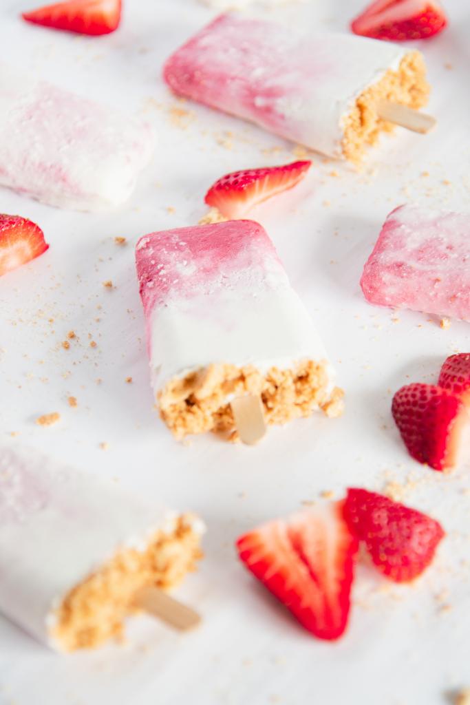 a close up of a cheesecake popsicle with chopped strawberries and graham cracker crumbs around it 