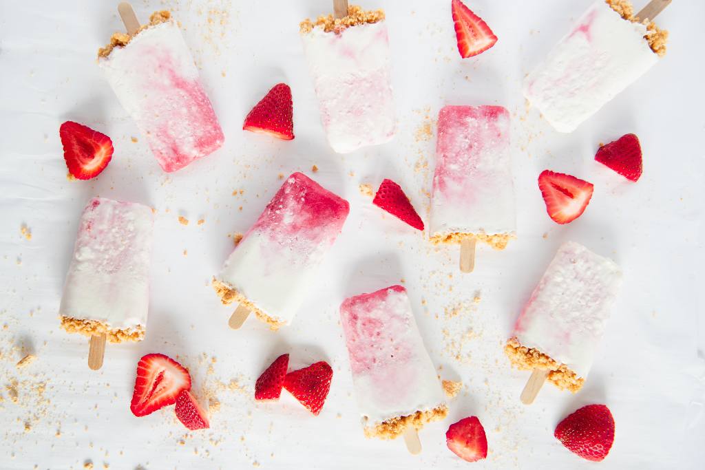 an overhead shot of a bunch cheesecake popsicles with chopped strawberries and graham cracker crumbs around them