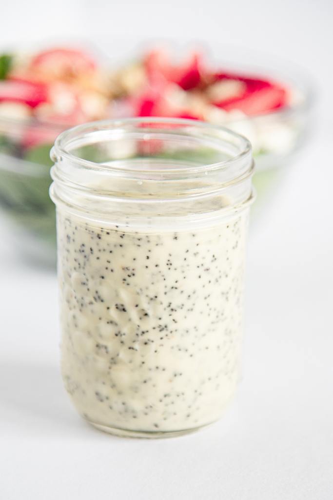 a close up of a jar of poppy seed dressing, you can see a bowl of salad behind it