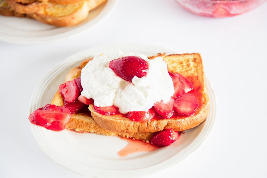 two pieces of french toast on a plate with strawberries and whipped cream on top, you can see a bowl of strawberries and another plate of french toast in the back ground 