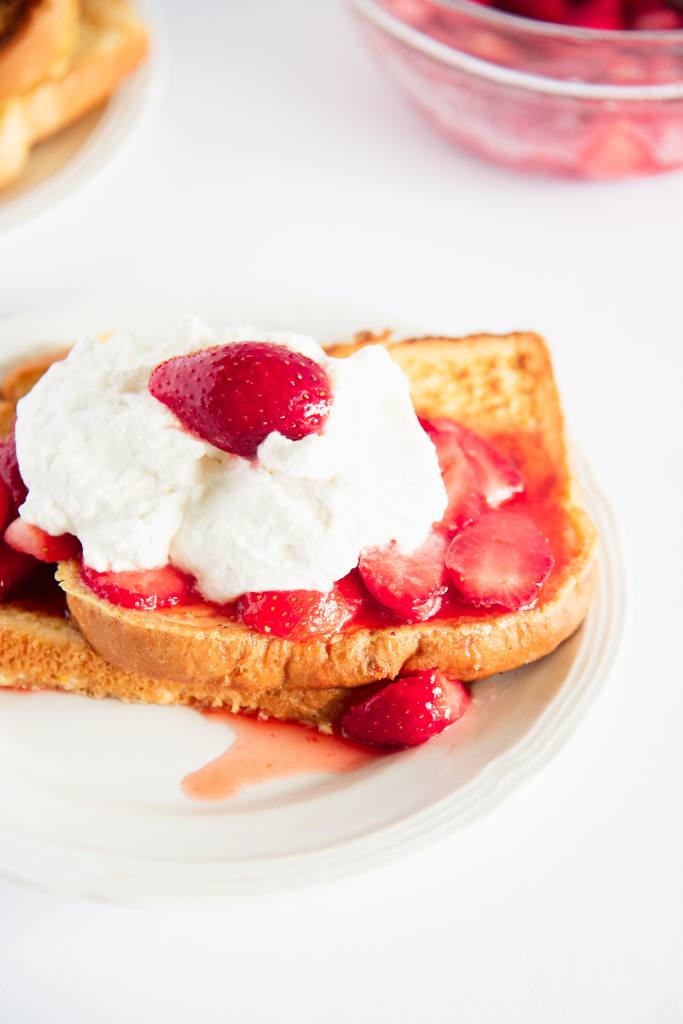 a cut of picture of a plate of french toast with strawberries and whipped cream on it, you can see part of a bowl of strawberries behind it 