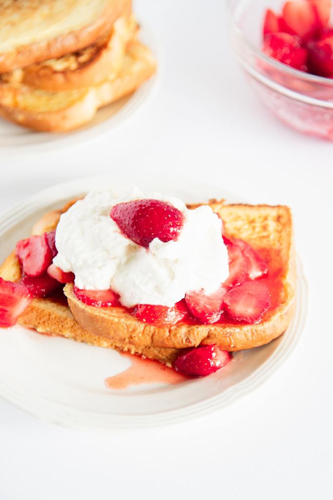 a plate of french toast with strawberries and whipped cream on top, you can see a bowl of strawberries and a plate of french toast behind it