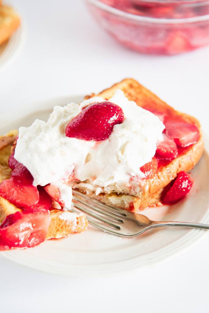 two pieces of french toast with strawberries and whipped cream on top, there is a bite taken out of it and a fork on the plate