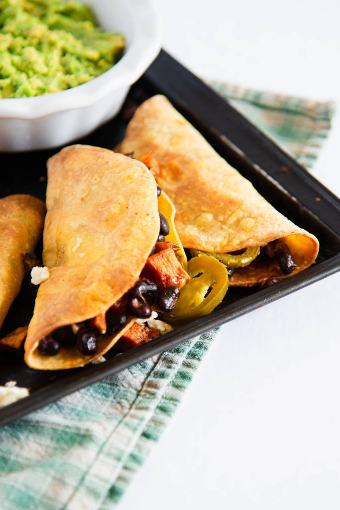 a cut off picture of a pan of crispy black bean and sweet potato tacos, there some jalapenos and a dish of guacamole on the pan as well