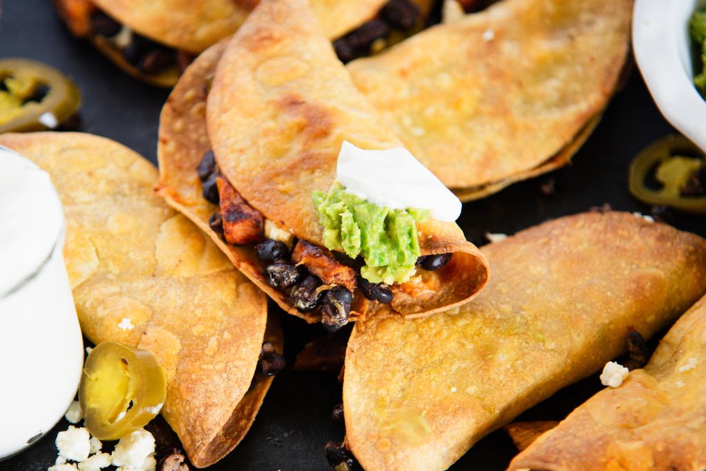 a pan of tacos with jalapenos around them, one taco has sour cream and guacamole a on it, and you can see part of a dish of sour cream and one of guacamole on either side of the pan