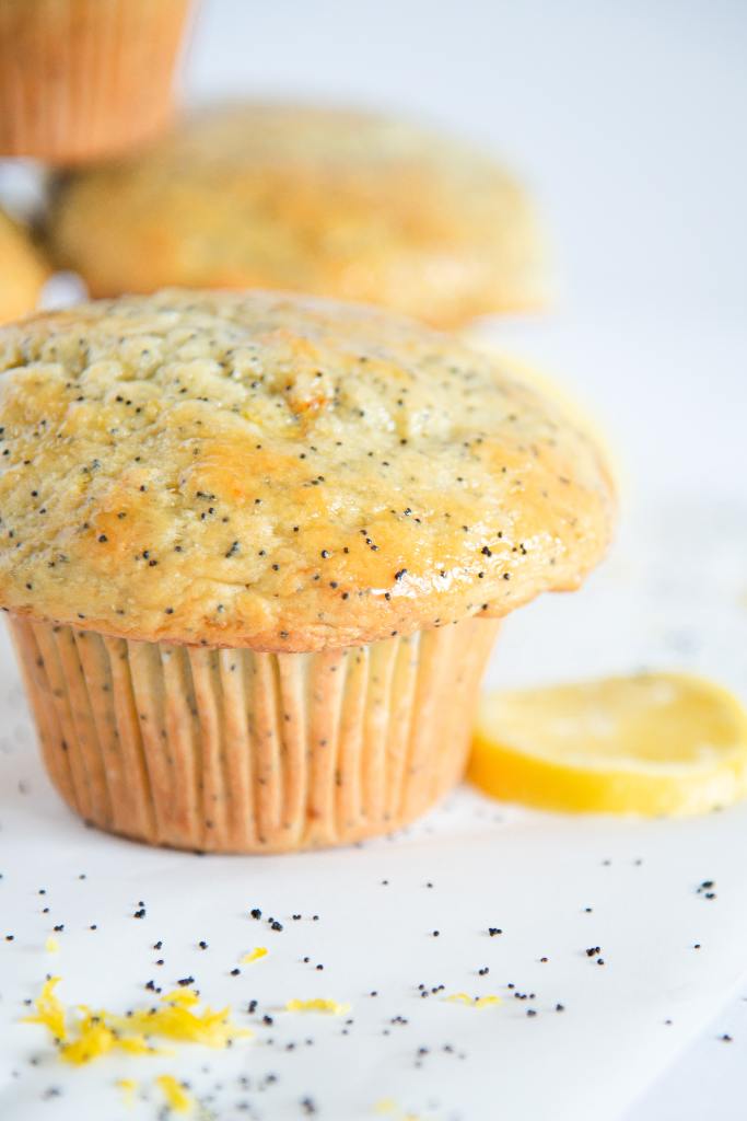 a close up of a lemon poppy seed muffin, you can see part of a lemon slice and some stacked muffins behind it