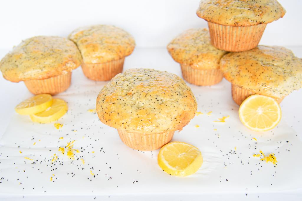 six lemon poppy seed muffins on a table, with lemon slices by them, and poppy seeds sprinkled around them.