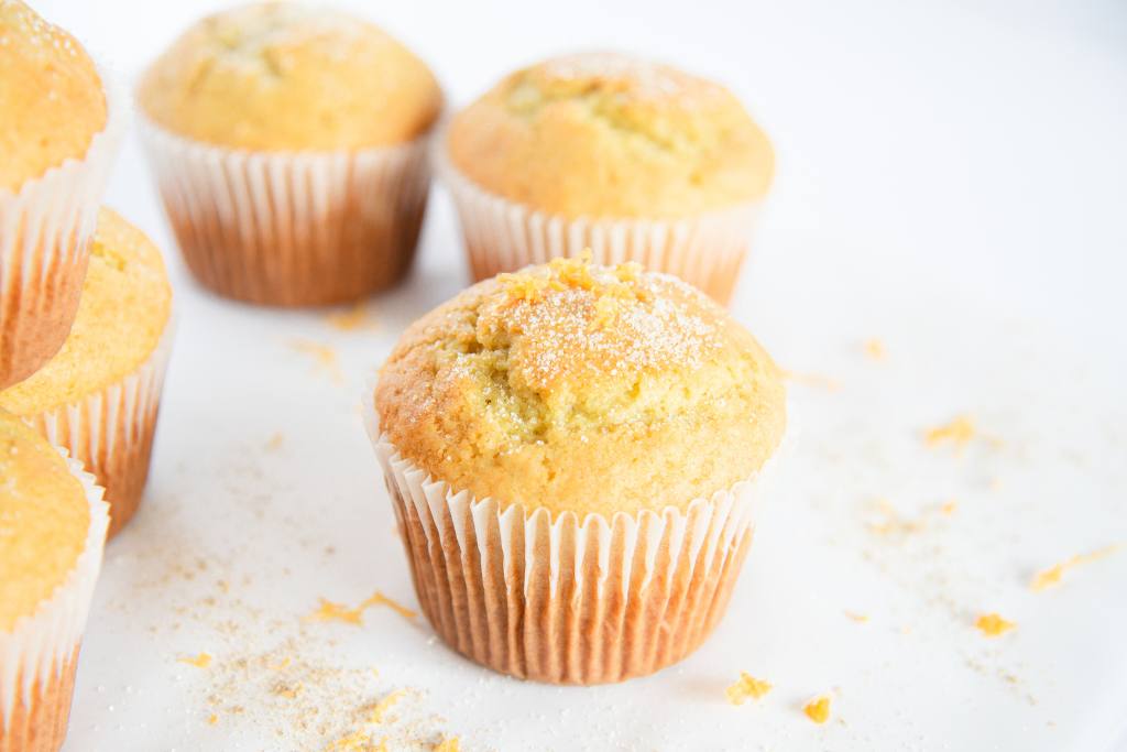 an orange cardamom muffin with sugar and orange zest sprinkled on top, are there some muffins stacked next to it and a couple muffins behind it