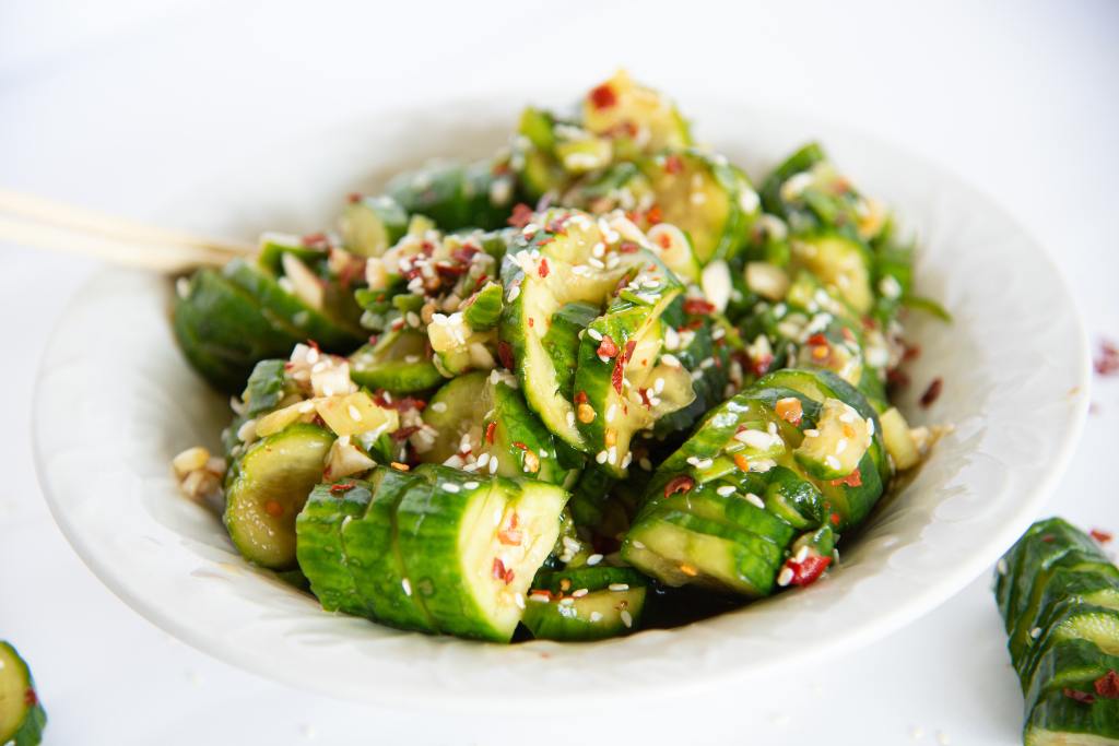 a bowl of spicy cucumbers with chopsticks stuck in it, there are some cucumber pieces on the table to the side of the bowl