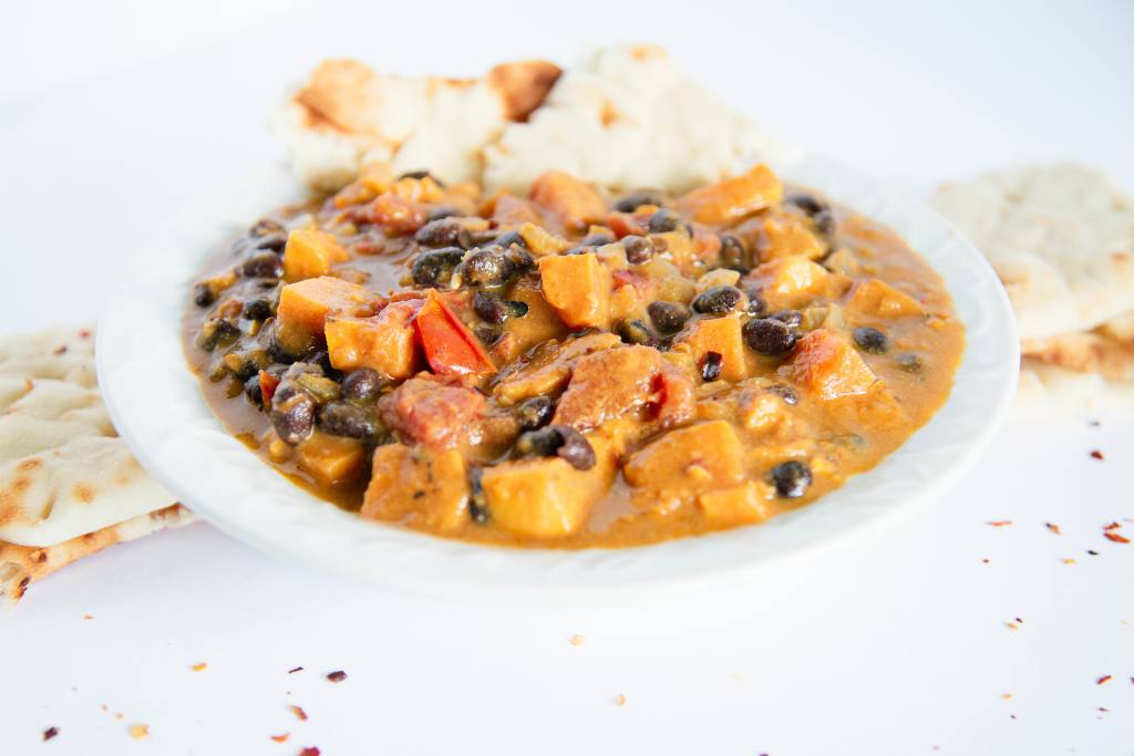 a bowl of black bean sweet potato curry with a piece of naan bread stuck in it, there are some pieces of naan bread next to the bowl as well