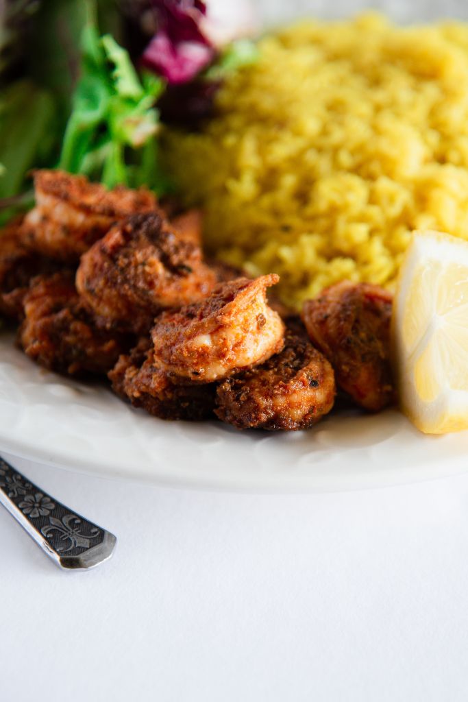 a close up of blackened shrimp on a plate, you can see some rice and greens behind it, and part of a lemon slice