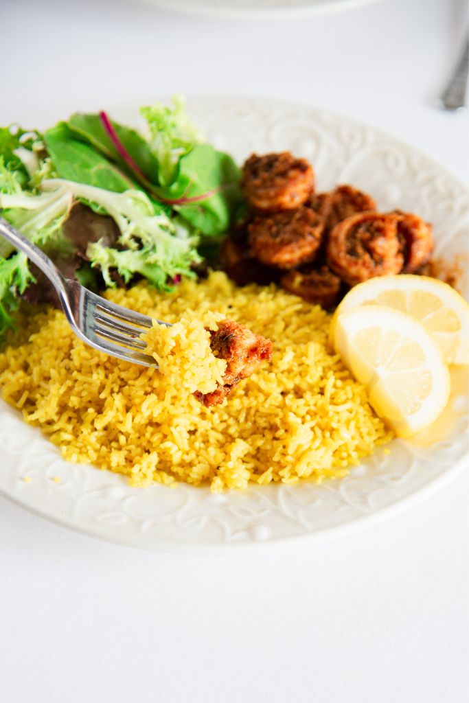 a fork with rice and a piece pf shrimp being held over a place of rice, shrimp and greens