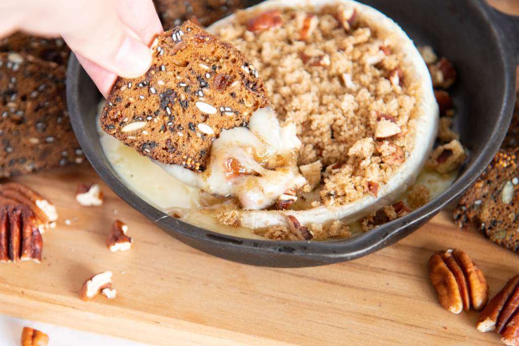 someone dipping a cracker into a wheel of baked brie. There are more crackers and some pecan pieces around the pan of brie