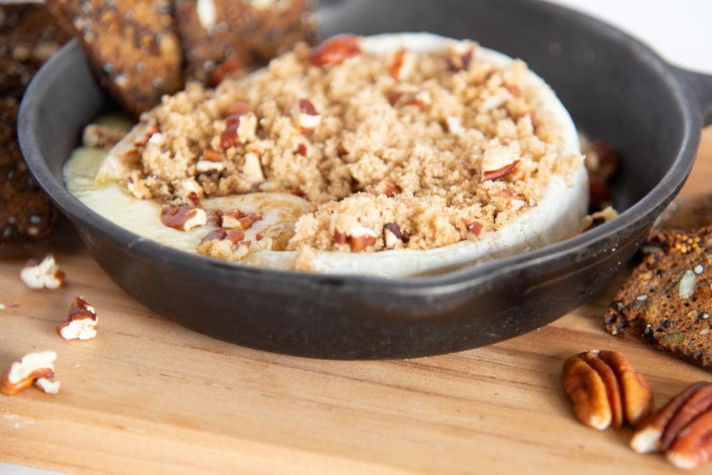 a close up of baked brie in a cast iron skillet, there is brown sugar and pecans on top of it, and some crackers around the pan