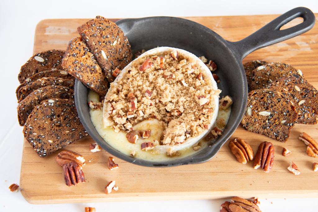 a wheel of baked brie that has been cut open in a cast iron skillet, there is brown sugar and pecans pieces on top the brie, and crackers and whole pecans around the pan